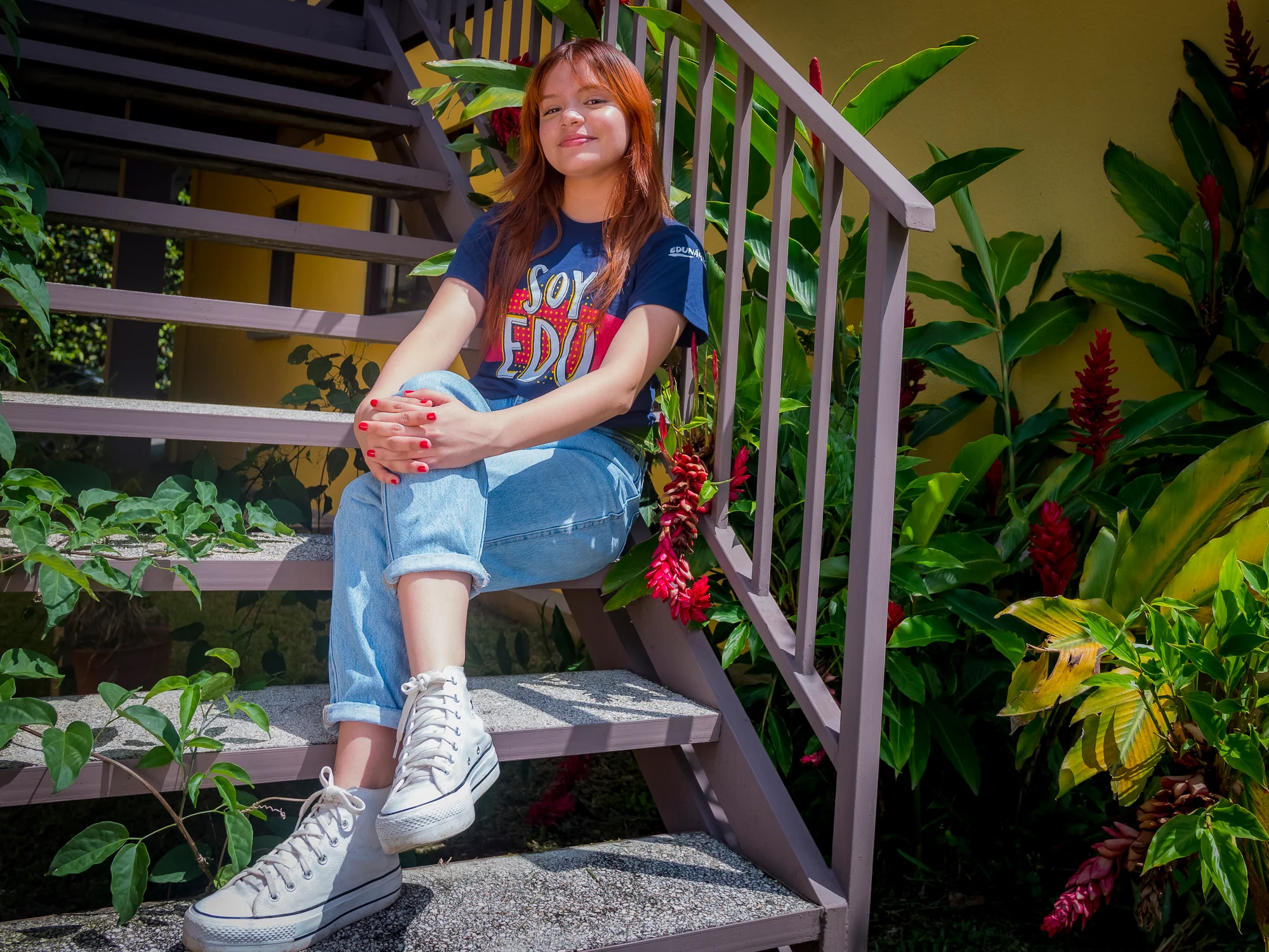 Edunámica beneficiary sitting on an outdoor staircase wearing the Soy Edunámica campaign t-shirt, surrounded by tropical plants