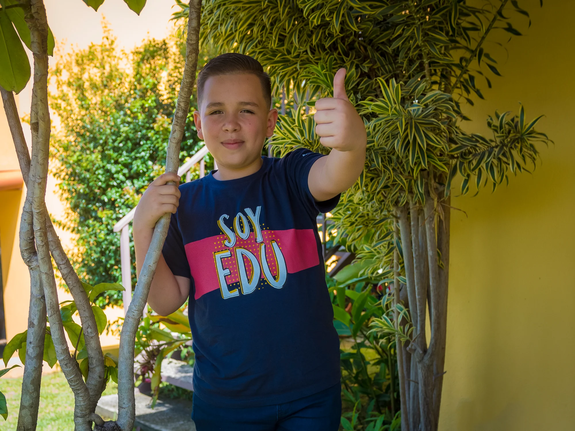 Edunámica beneficiary wearing a Soy Edunámica t-shirt giving a thumbs up among garden foliage