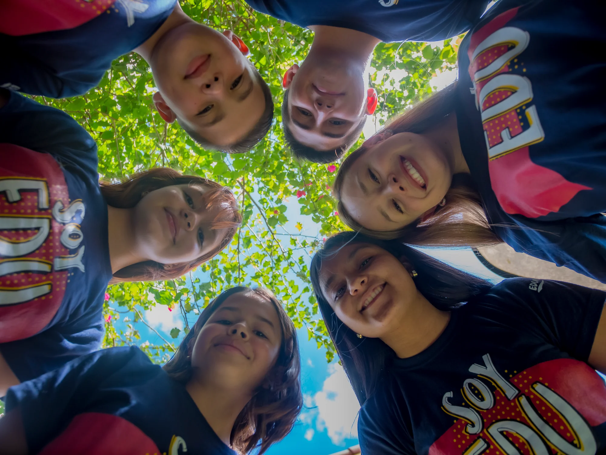 Group of Edunámica beneficiaries in a circle, looking down at the camera with trees and sky in the background