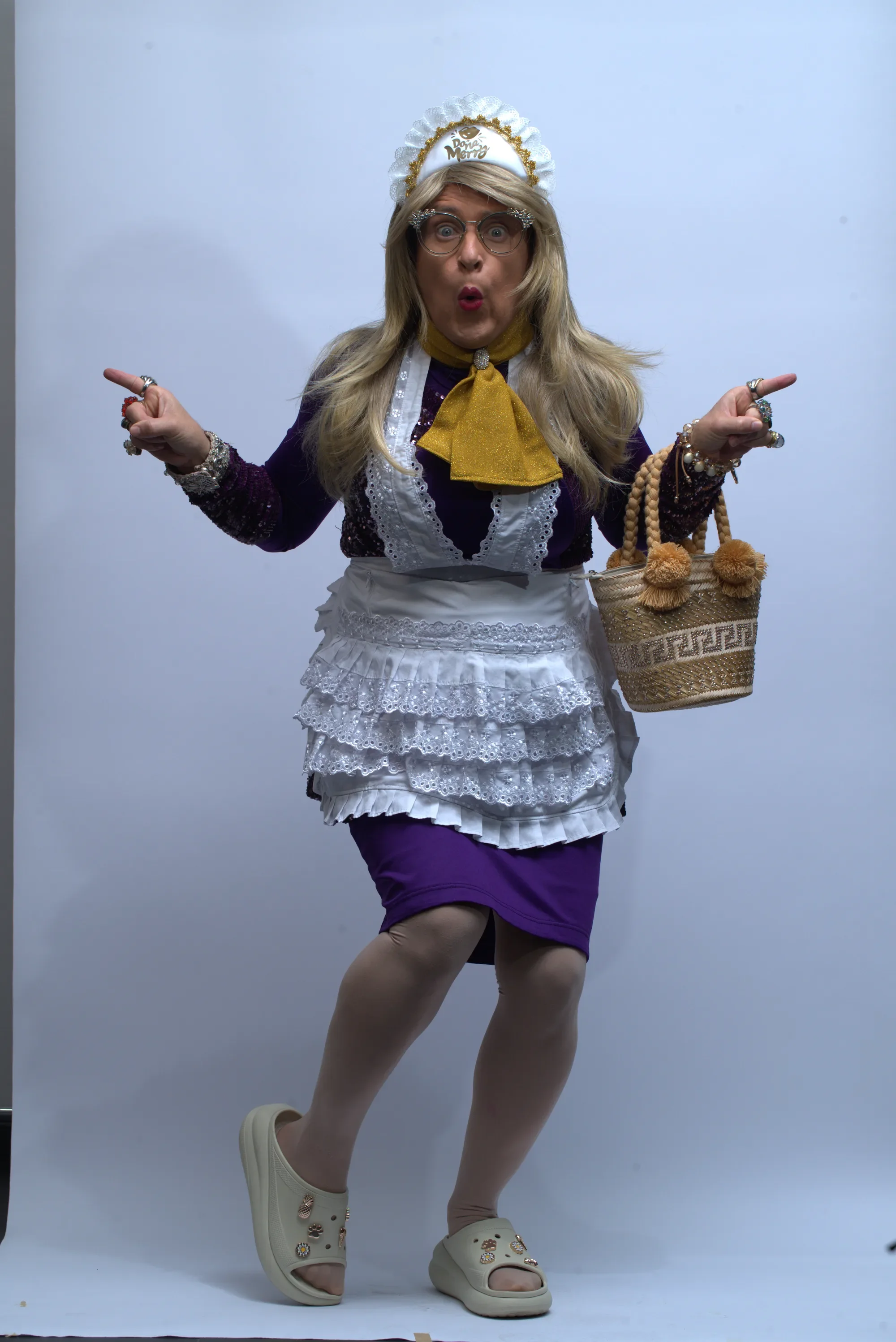 Portrait of Doña Merry pointing at the camera, wearing a lace apron and carrying a wicker purse